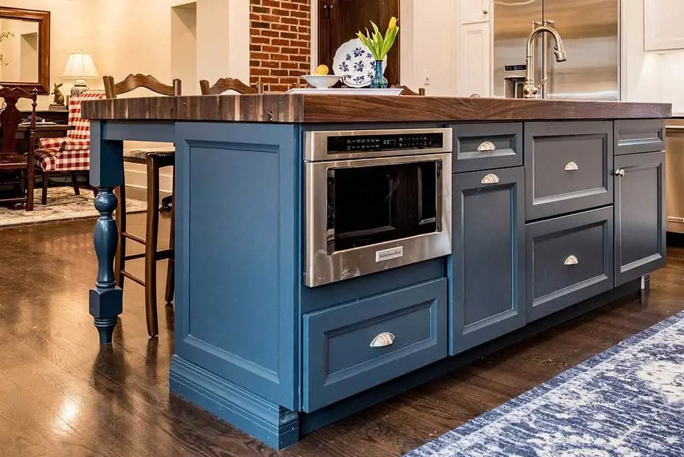 Blue kitchen island with a built-in microwave installed by appliance technicians, wooden countertop, dark floors, and a vase with flowers. Dining room with red-checkered chairs visible in the background.