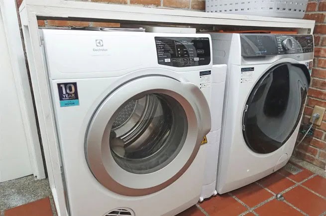 A front-loading washing machine and dryer sit side by side against a brick wall, with a white shelf and laundry basket above—ideal for those seeking MA appliance services or quick same day appliance repair.
