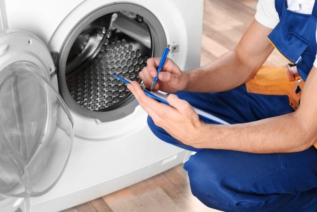 A repair technician in blue overalls kneels beside an open washing machine in Middlesex County, writing notes on a clipboard while inspecting the appliance for repairs. Same day appliance repair services are available.