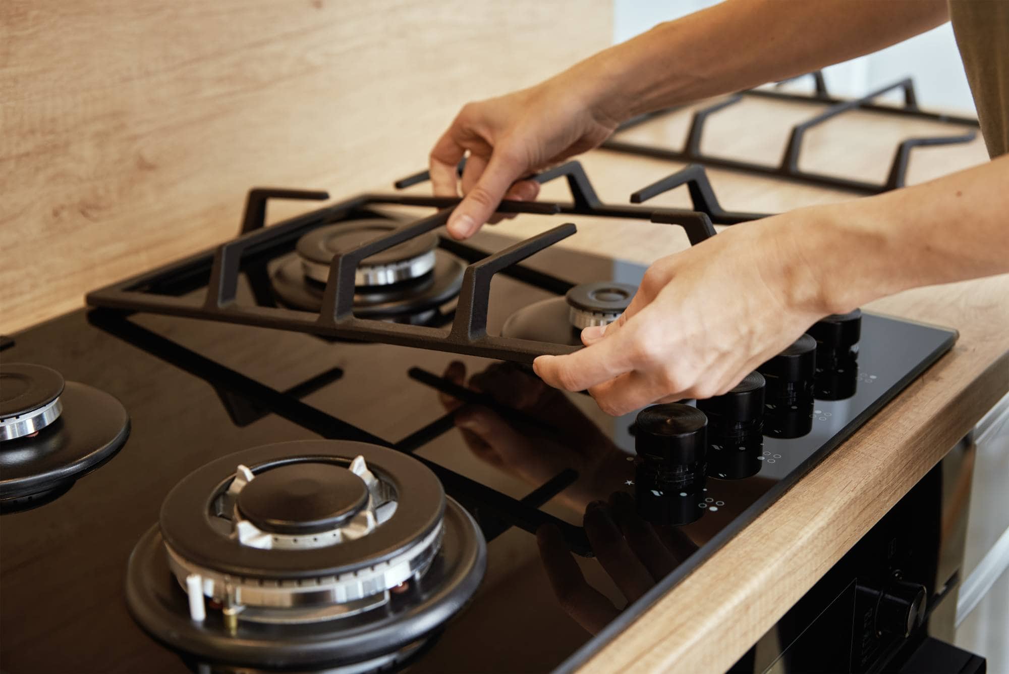 Close-up of a person’s hands placing a metal grate onto a black gas stovetop with multiple burners in a modern kitchen—just the kind of scene appliance technicians from MA appliance services handle daily in Middlesex County.