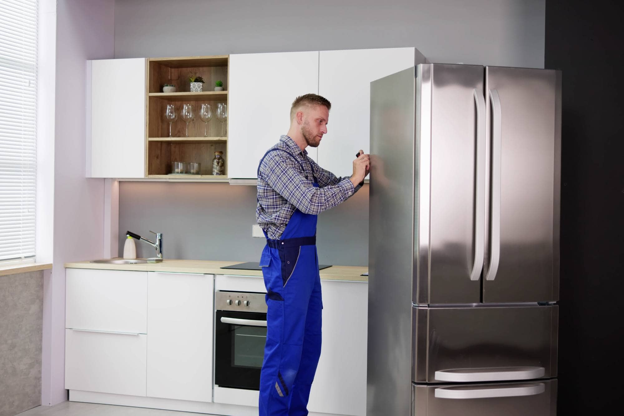 A repairman in blue overalls, one of the skilled appliance technicians in Middlesex County, is fixing a stainless steel refrigerator in a modern white kitchen with wooden accents and open shelving above the sink.