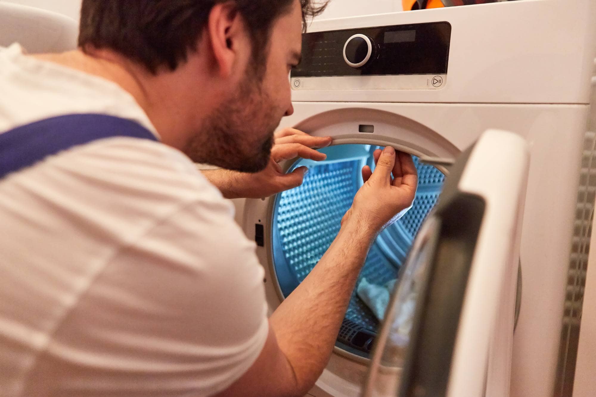 A man wearing a white shirt and blue overalls is inspecting or repairing the door seal of a front-loading washing machine, similar to skilled appliance technicians in Middlesex County who offer same day appliance repair.