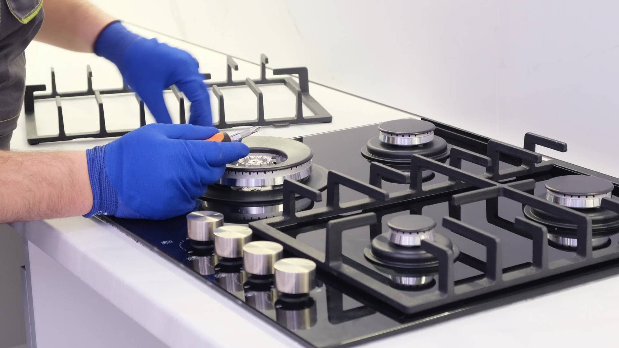 A person wearing blue gloves uses a screwdriver to repair or install a gas stove burner on a modern black glass cooktop, demonstrating the skilled work of appliance technicians in Middlesex County.