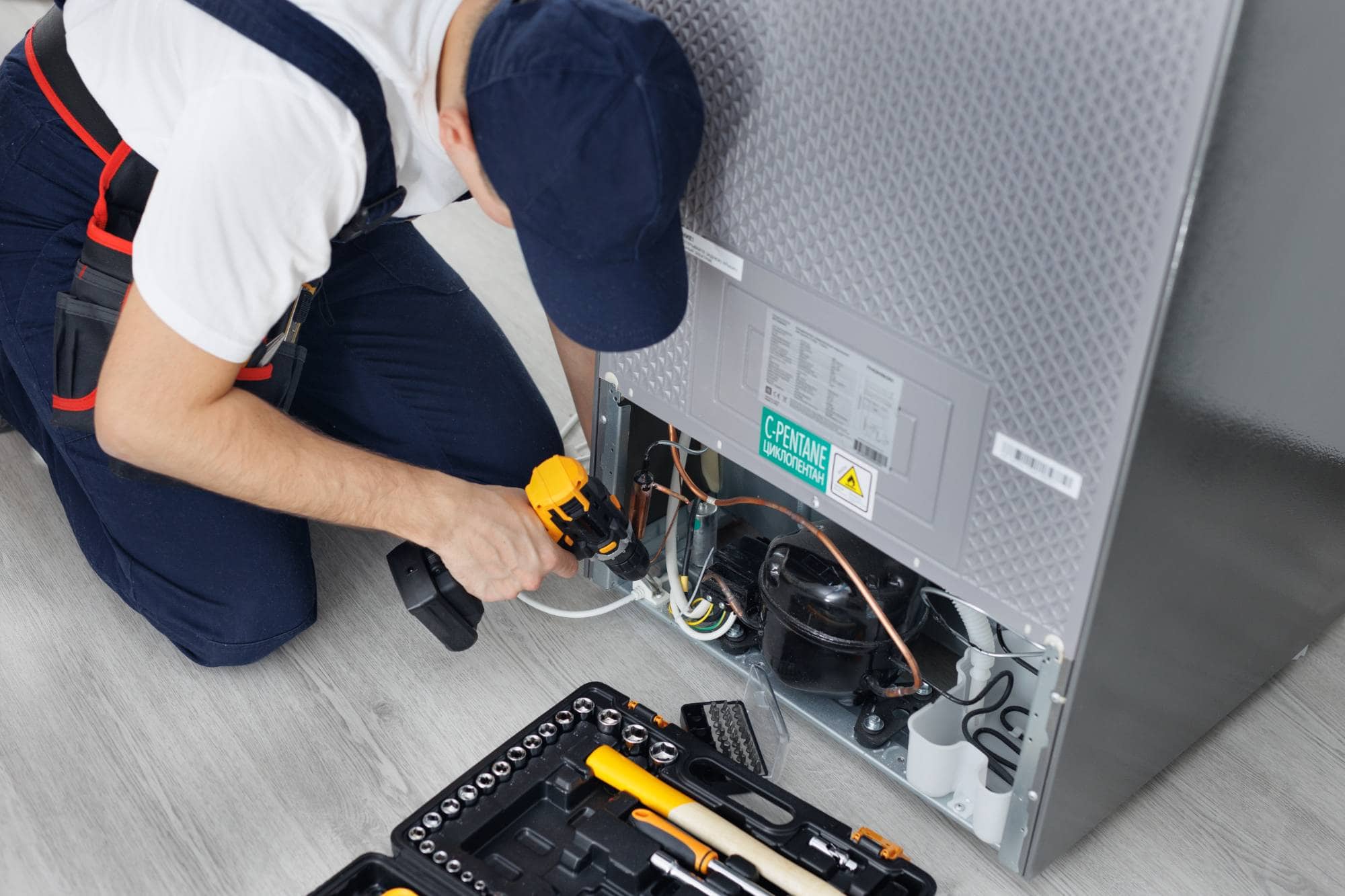 A technician in uniform uses a cordless drill to repair the lower back panel of a refrigerator. For prompt Middlesex County, MA appliance services, his toolkit is laid out on the floor, ready for same day appliance repair.