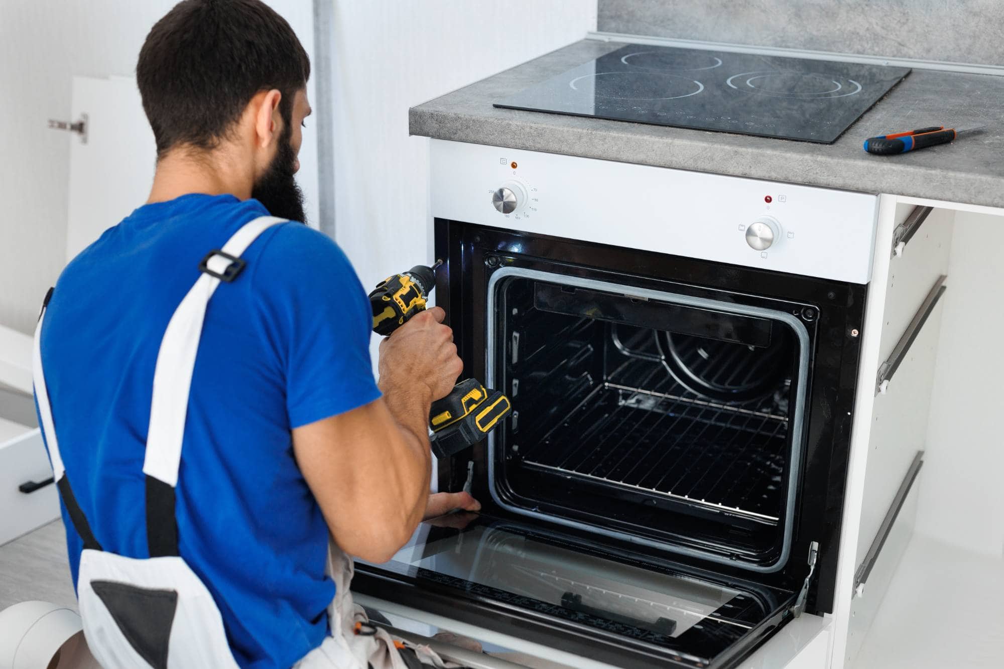 A man in blue overalls uses a cordless drill to repair or install a built-in oven in a modern kitchen. The oven door is open, and a screwdriver rests on the countertop above—showcasing expert MA appliance services.