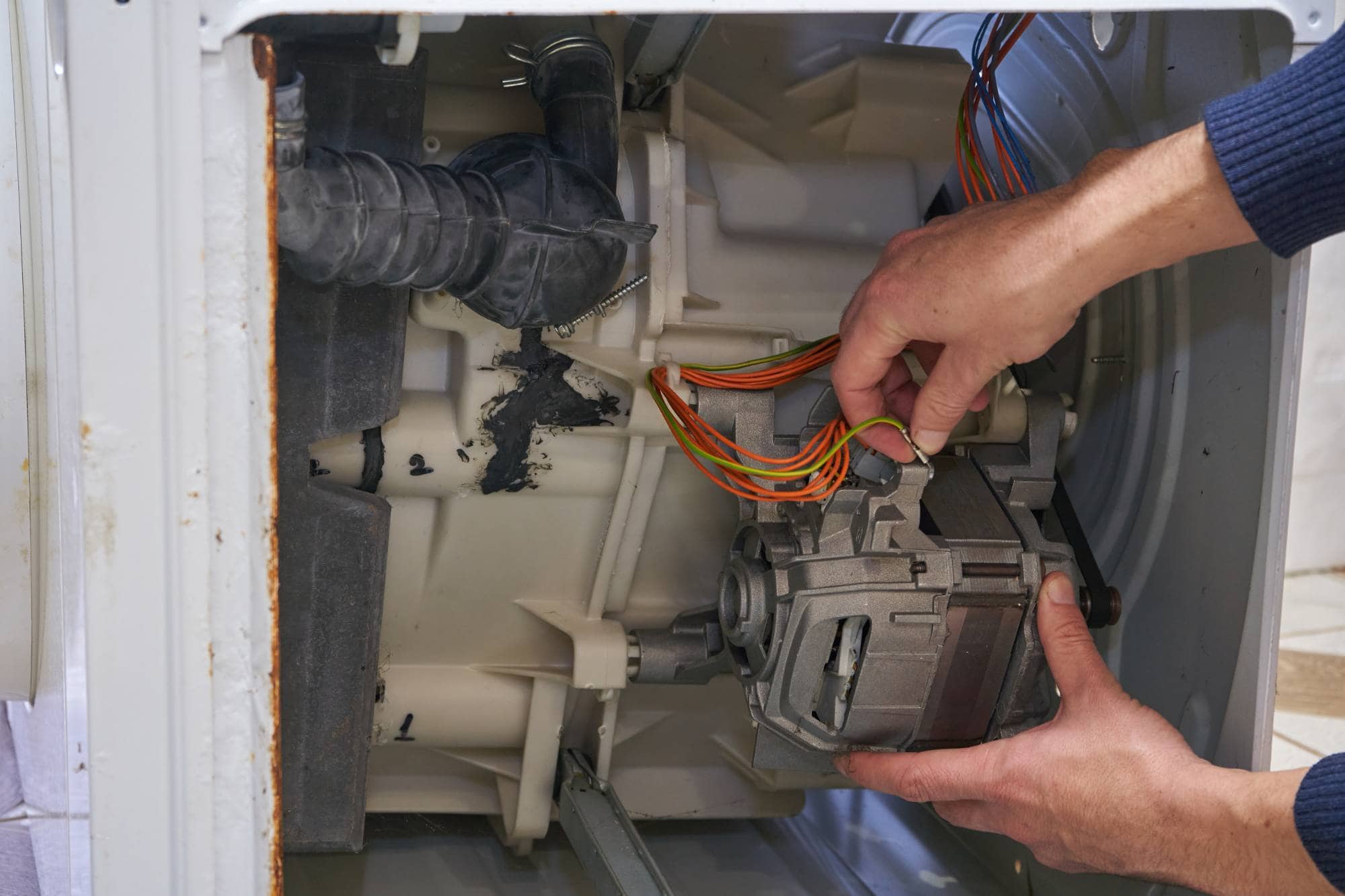 A person provides same day appliance repair, skillfully holding colorful wires and using both hands to adjust components inside the open back panel of a washing machine.