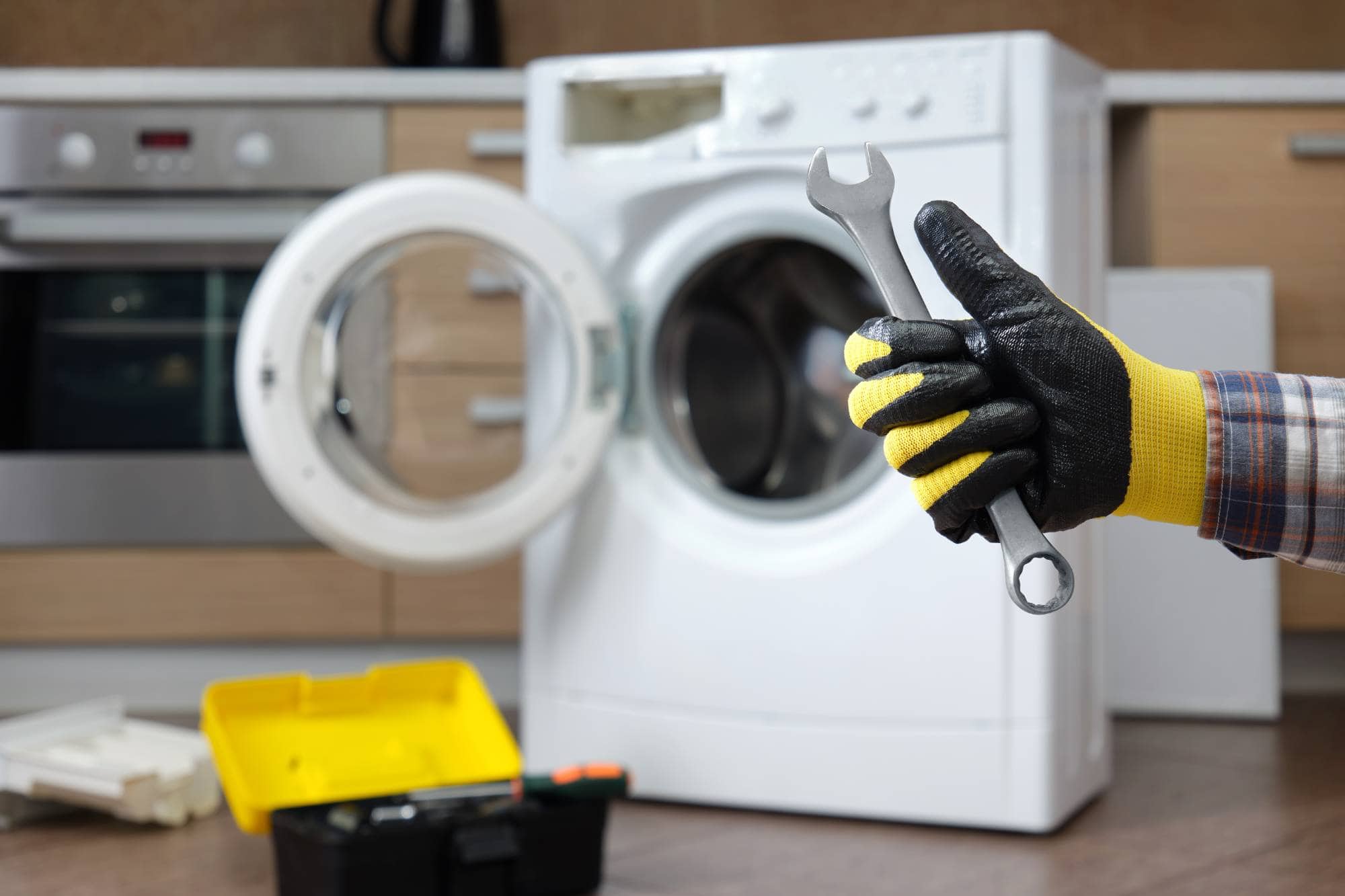 A person wearing work gloves holds a wrench in front of an open washing machine, with a toolbox on the floor—showcasing same day appliance repair in a modern kitchen or laundry area.