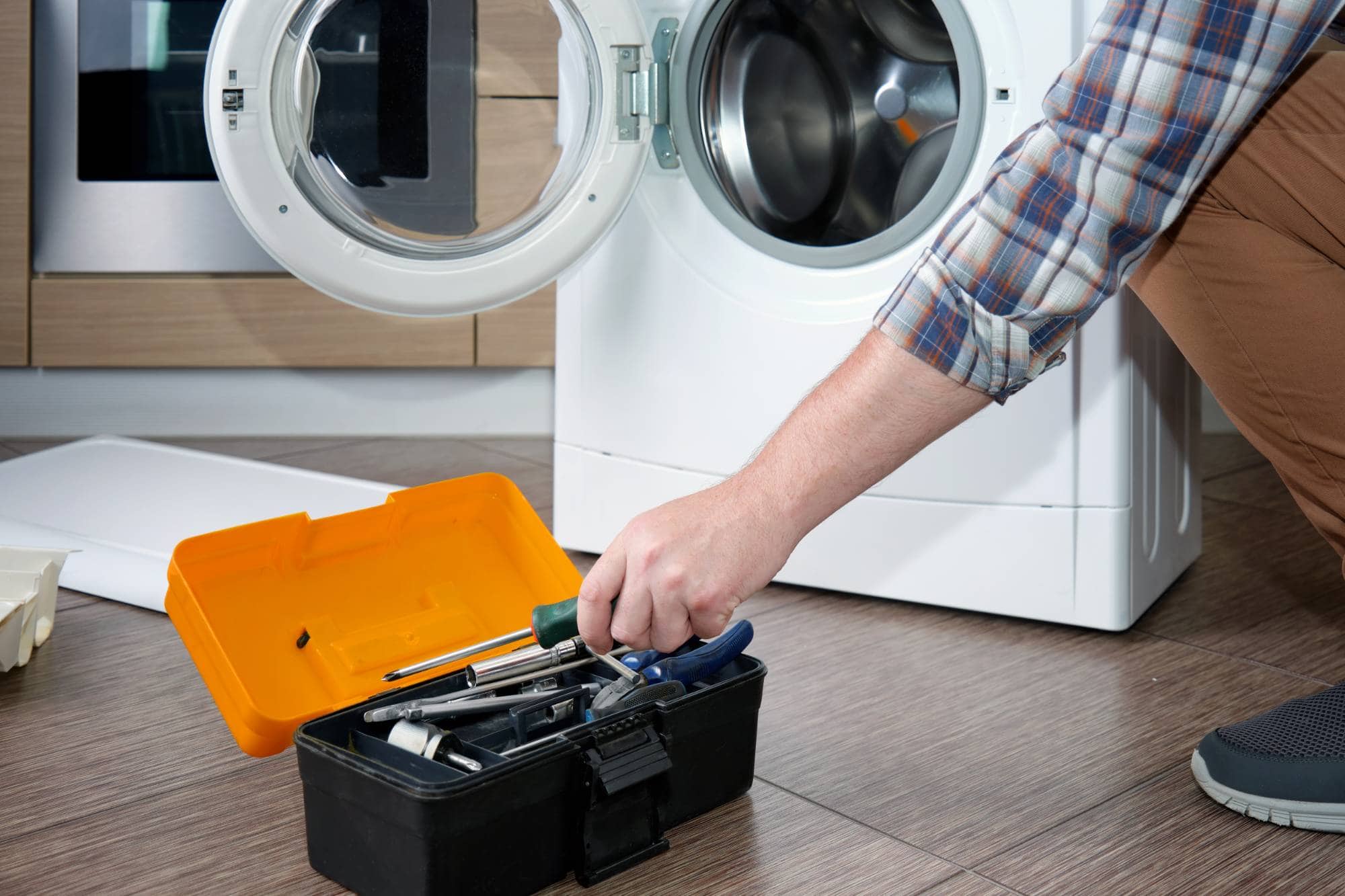 A person kneels beside an open washing machine in a modern laundry room, reaching into a black toolbox with an orange lid—illustrating appliance technicians at work, possibly providing MA appliance services in Middlesex County.