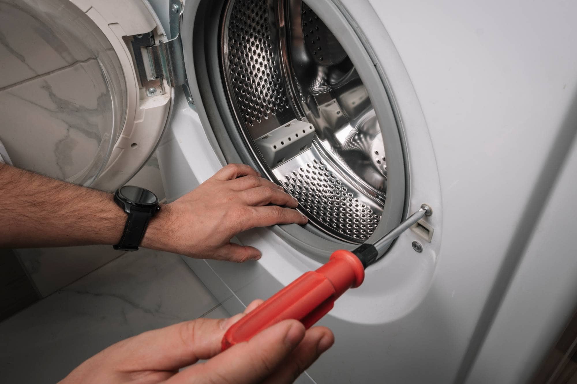A person uses a red screwdriver to repair or inspect the open door seal area of a front-loading washing machine, demonstrating the expertise of Middlesex County appliance technicians. One hand holds the screwdriver while the other steadies the door.