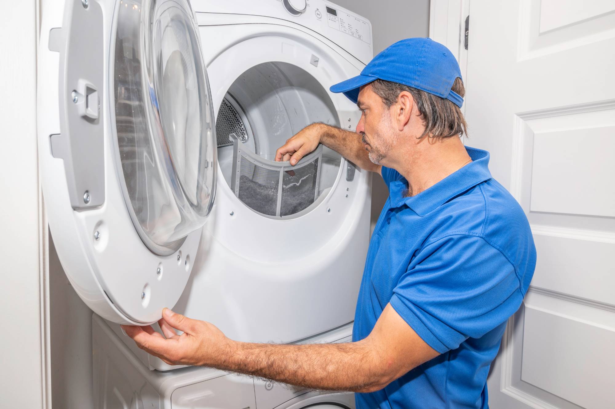 A man in a blue shirt and cap is cleaning the lint filter of a front-loading dryer in a Middlesex County laundry room. The dryer door is open as he removes debris, highlighting the importance of regular MA appliance services.