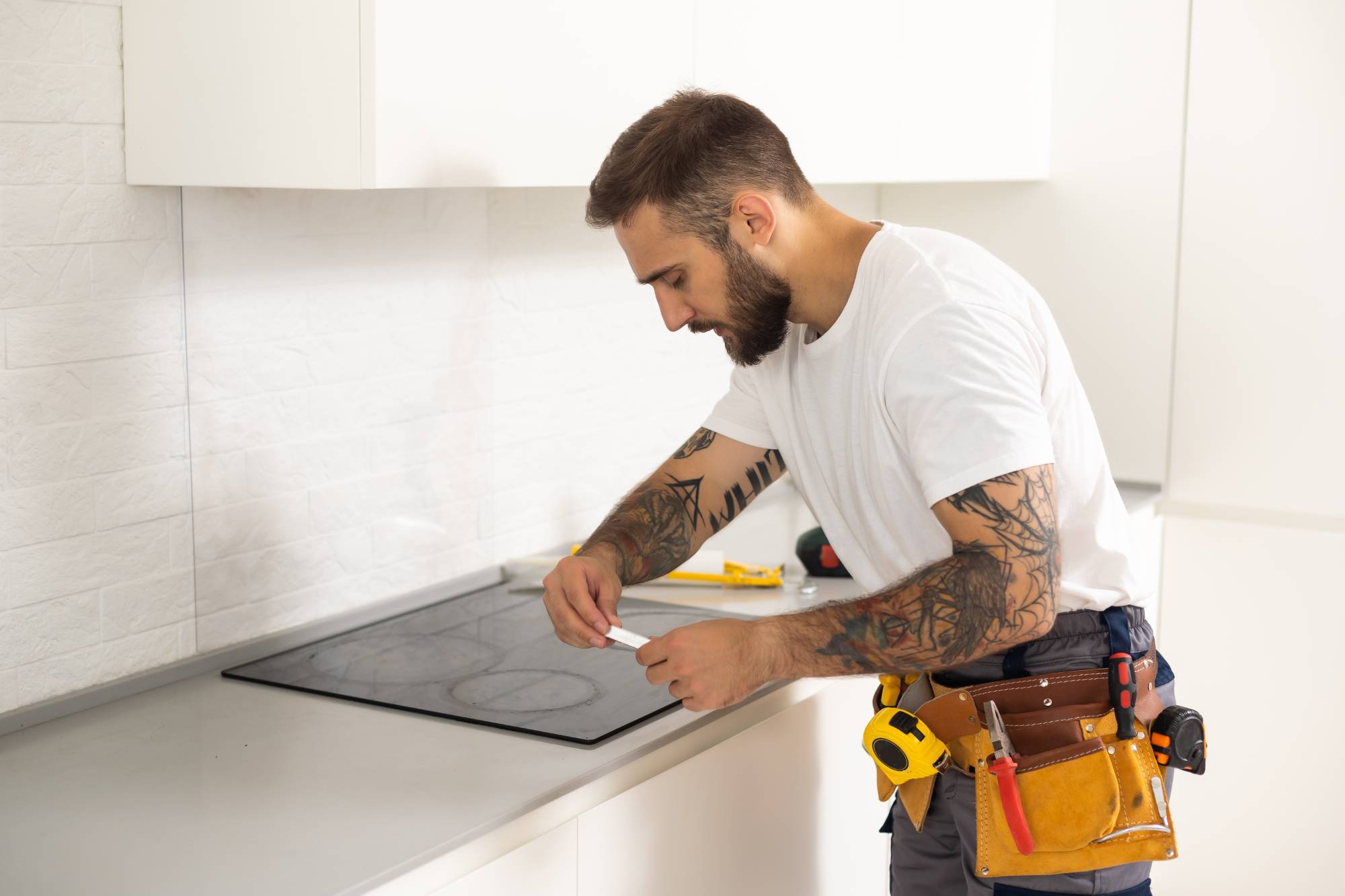 A bearded man with tattoos, wearing a white t-shirt and tool belt, installs or repairs a glass cooktop in a modern Middlesex County kitchen. Appliance technicians' tools are visible on the counter.