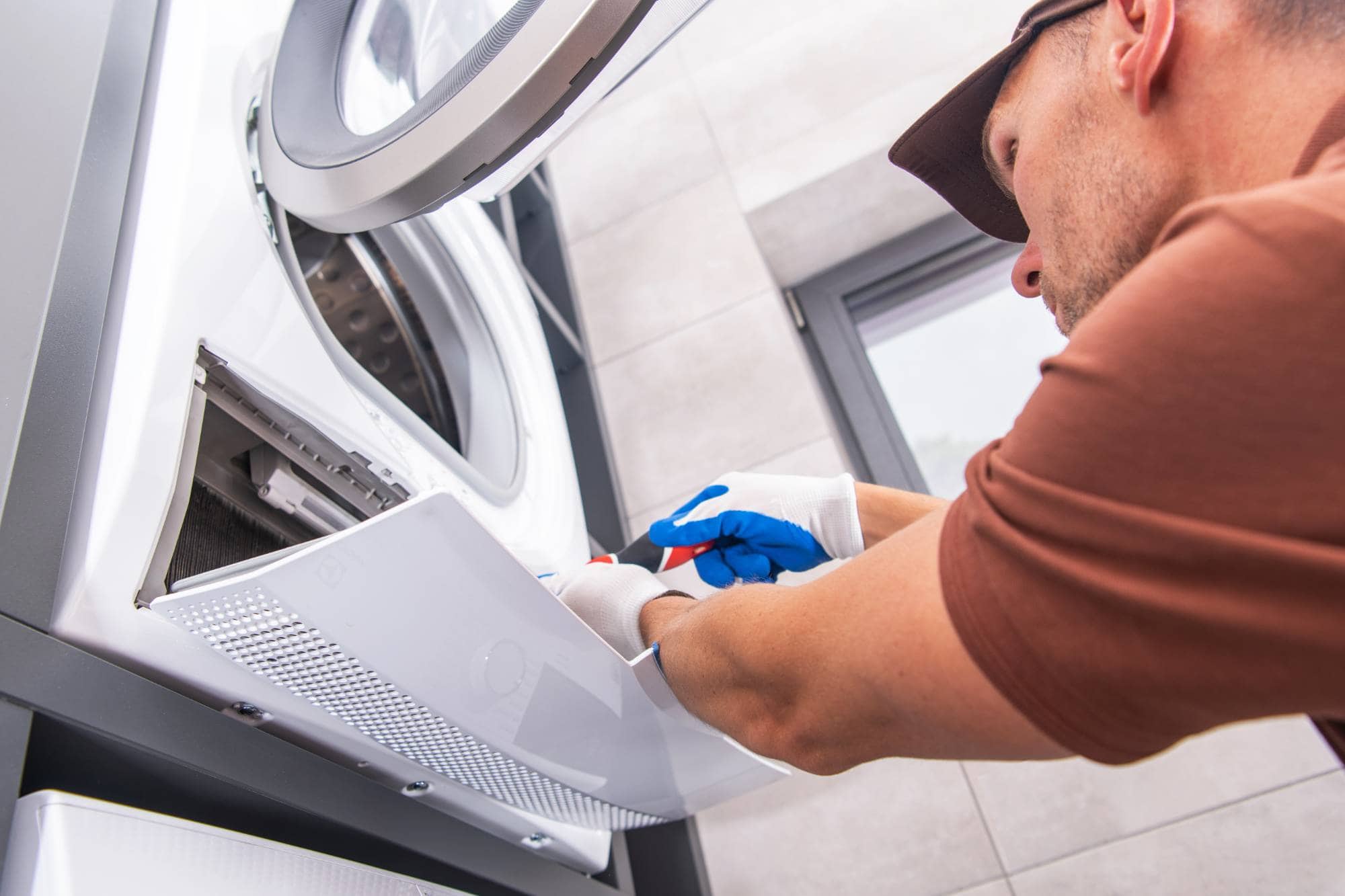 A man wearing gloves and a cap is repairing or inspecting the front panel of a washing machine in a bright, tiled room. As one of Middlesex County’s expert appliance technicians, he uses tools to ensure quality same day appliance repair.