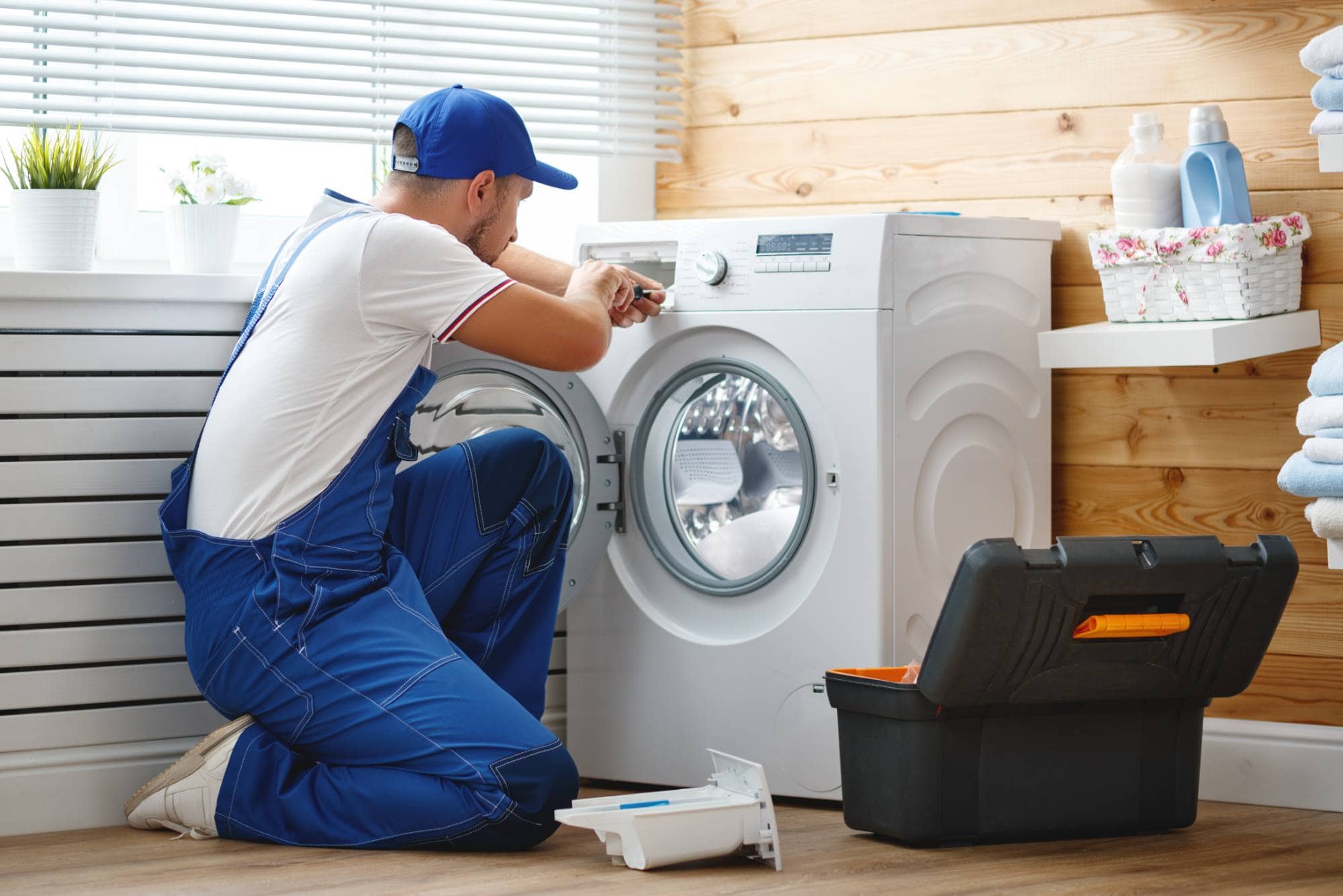 A repairman in blue overalls kneels in front of a washing machine, using a tool to fix it. A toolbox is open on the floor nearby. The bright laundry room shows shelves, towels, and potted plants—perfect for same day appliance repair in Middlesex County.