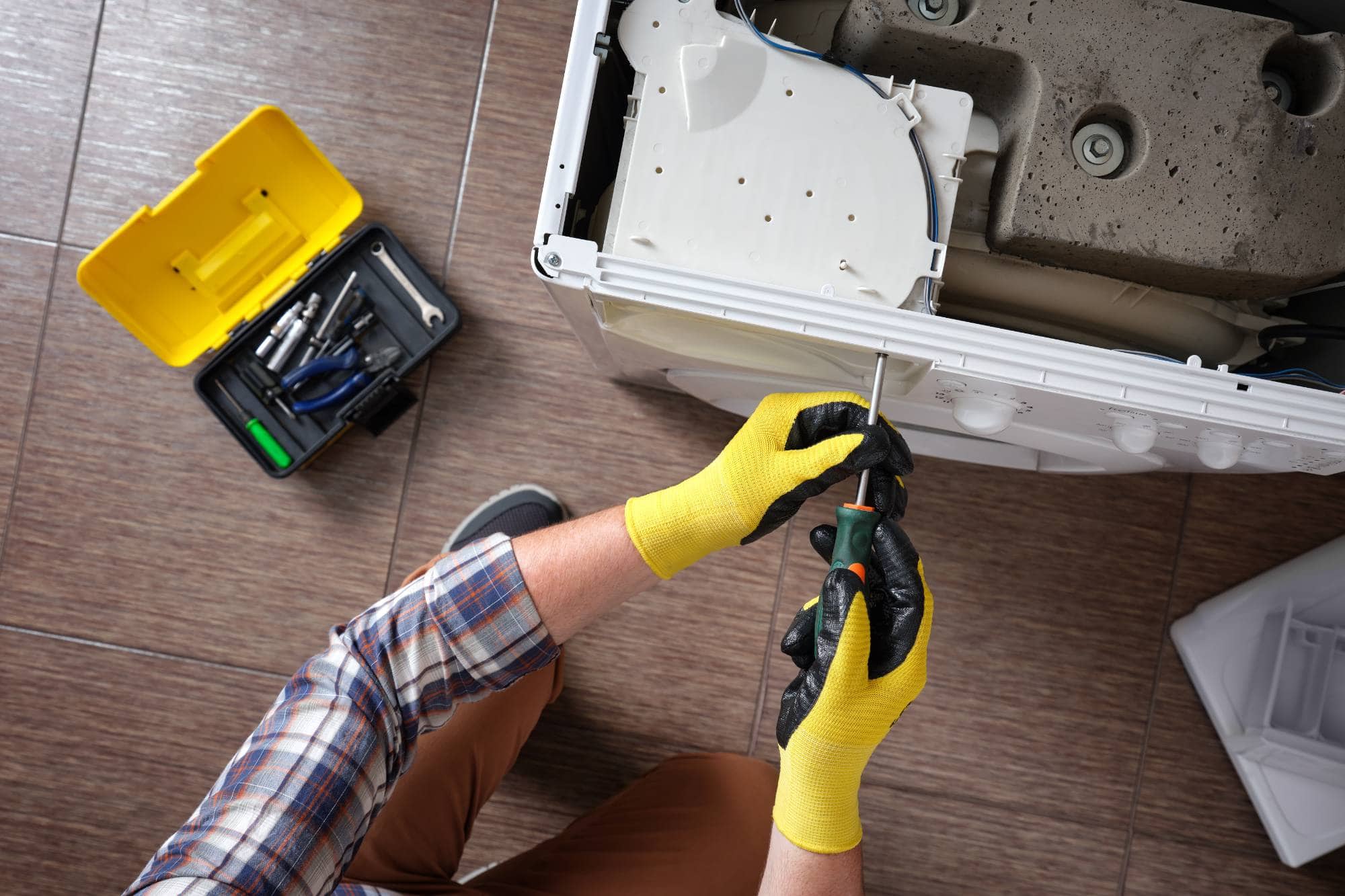 A person wearing yellow gloves uses a screwdriver to repair a washing machine, with a toolbox full of tools on the tiled floor nearby—showcasing the expert touch of MA appliance services for reliable repairs for appliances.