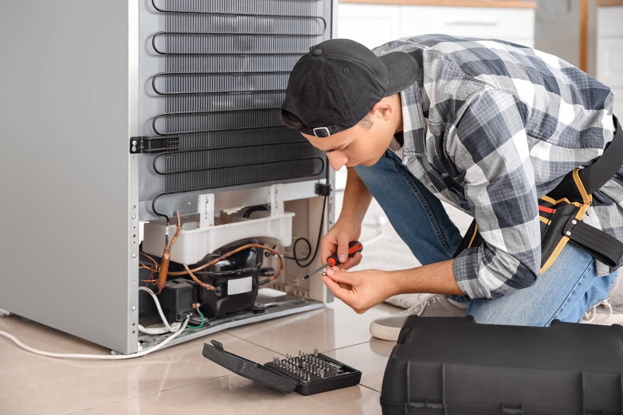 A man in a plaid shirt and backwards cap kneels on the floor using pliers to repair the back of a refrigerator, with a tool kit open beside him—demonstrating the skill of professional appliance technicians handling repairs for appliances.