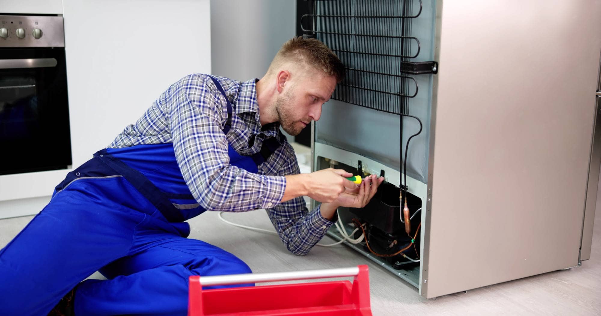 A repairman in blue overalls, representing MA appliance services, is kneeling and fixing the back of a refrigerator with a screwdriver in a modern kitchen. A red toolbox sits on the floor nearby, showing dedication to quality repairs for appliances.