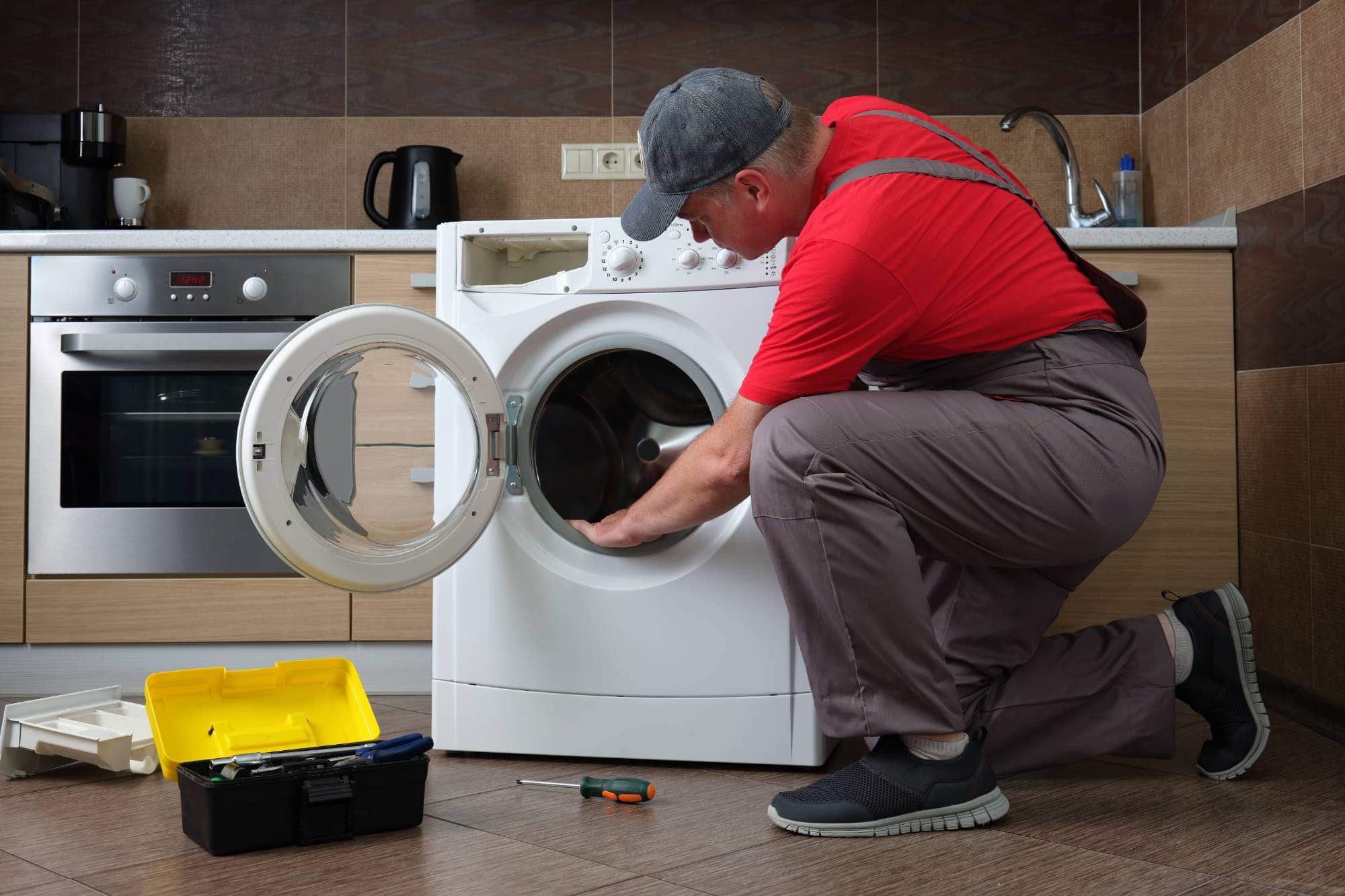 A repairman in a red shirt and gray cap kneels in front of an open washing machine, inspecting its drum. A yellow toolbox and tools are on the floor beside him—a typical scene for appliance technicians handling same day appliance repair in a modern kitchen.