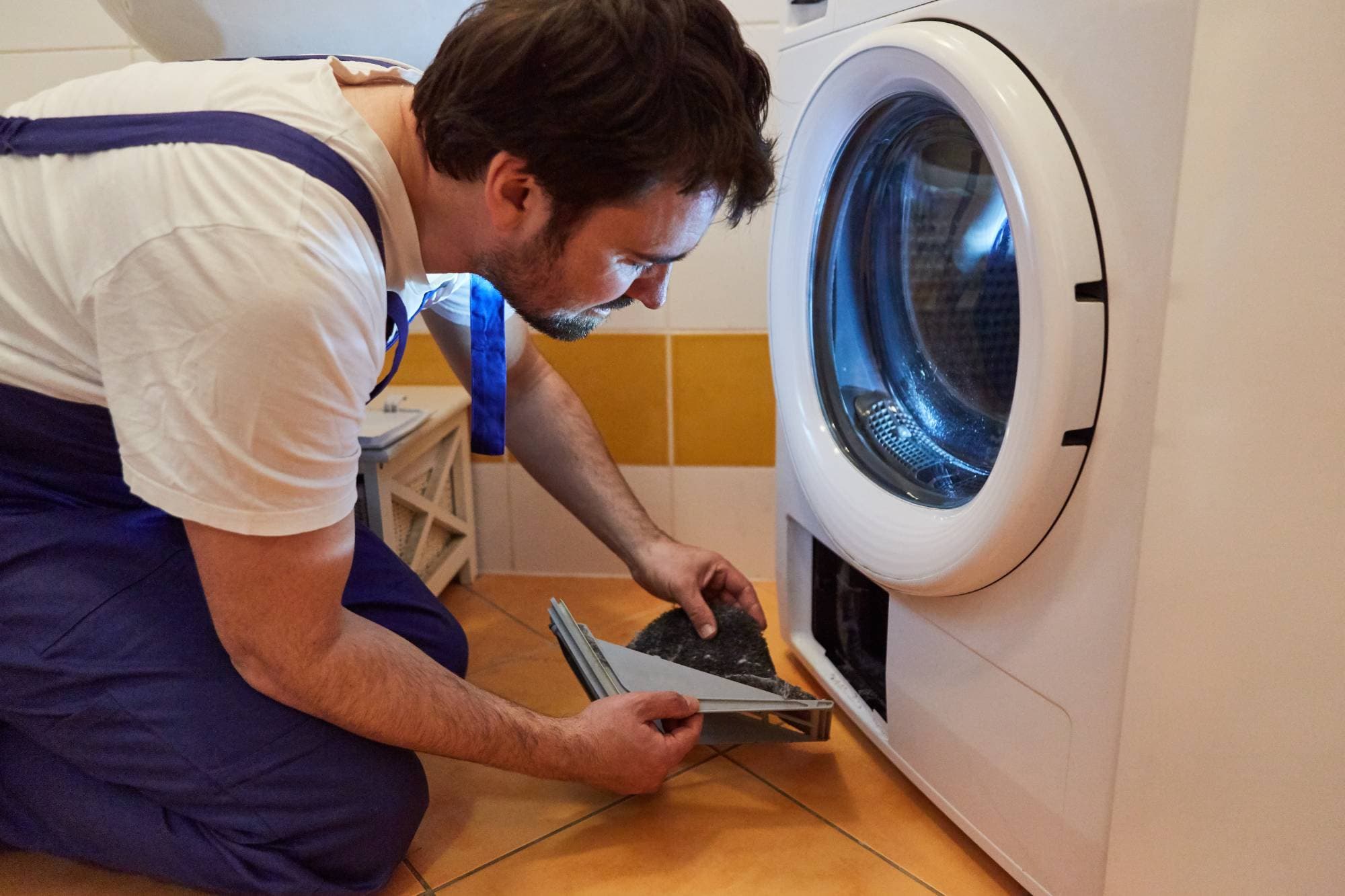 A man in work overalls kneels on the floor while cleaning or repairing the filter compartment of a front-loading washing machine, showcasing skilled appliance technicians at work in a tiled laundry room.