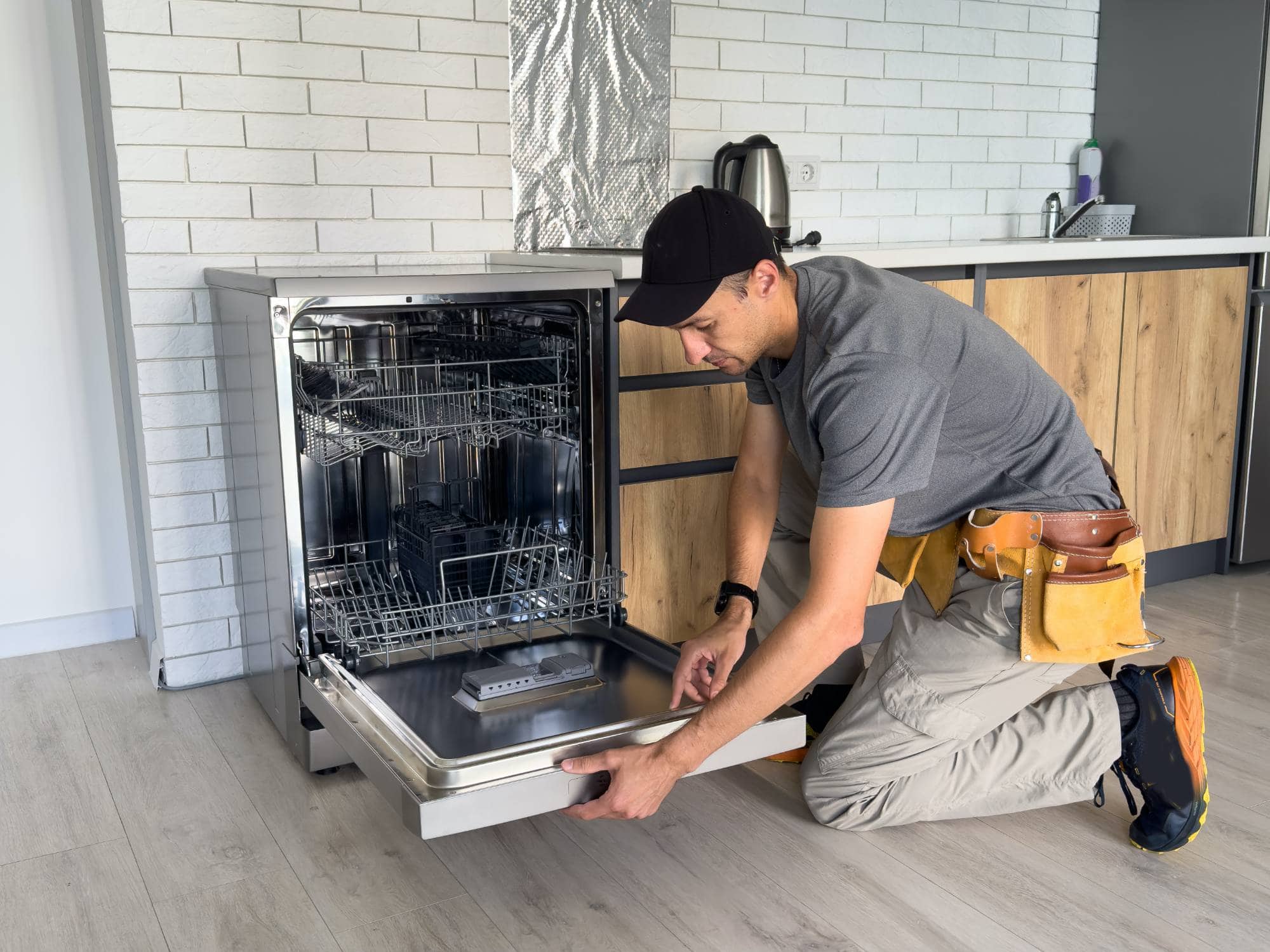 A repairman wearing a tool belt kneels on the floor while working on an open dishwasher in a modern Middlesex County kitchen, showcasing the skills of experienced appliance technicians among wooden cabinets and white brick walls.