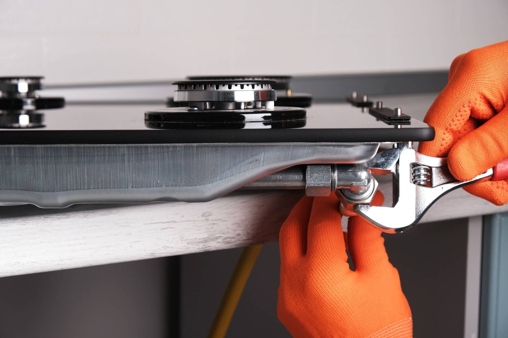 A person wearing orange gloves uses a wrench to tighten a gas connection under a stovetop during installation or maintenance, just like the expert appliance technicians providing same day appliance repair in Middlesex County.