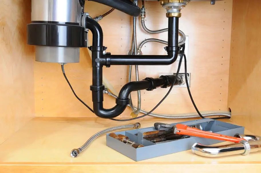 Plumbing pipes and a garbage disposal unit under a kitchen sink, with a tray of wrenches and pliers on the cabinet floor—ready for MA appliance services or same day appliance repair in Middlesex County.