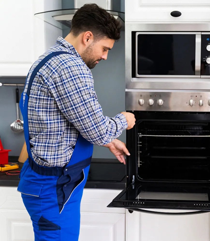 A man in blue overalls and a plaid shirt is opening the door of a built-in kitchen oven, appearing to inspect or repair it—just like skilled appliance technicians offering same day appliance repair in a modern kitchen with white cabinets and a microwave above the oven.