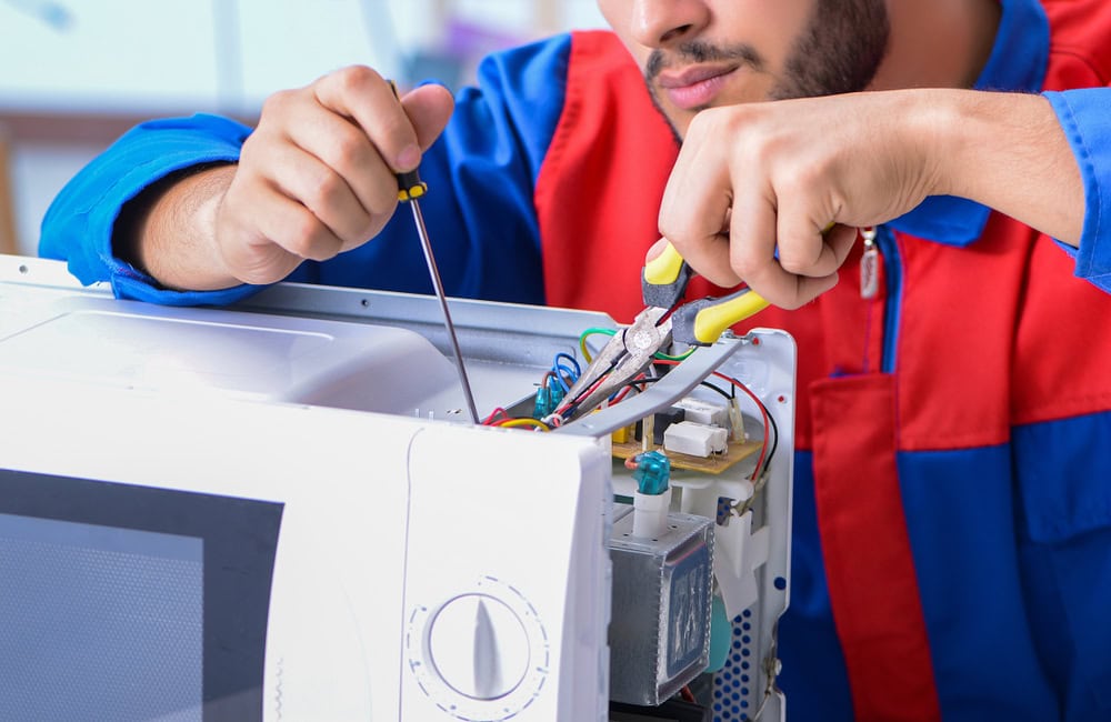 A person in a red and blue uniform, one of the skilled appliance technicians, is repairing a microwave oven, using a screwdriver and pliers to work on the internal wiring and components.