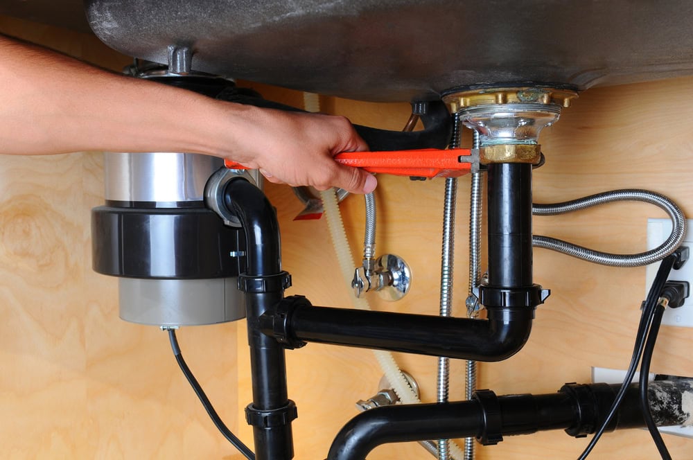 A person uses an orange wrench to tighten or loosen a pipe connection under a sink, showing black plumbing pipes and a garbage disposal unit—demonstrating repairs for appliances performed by MA appliance services against a wooden cabinet background.