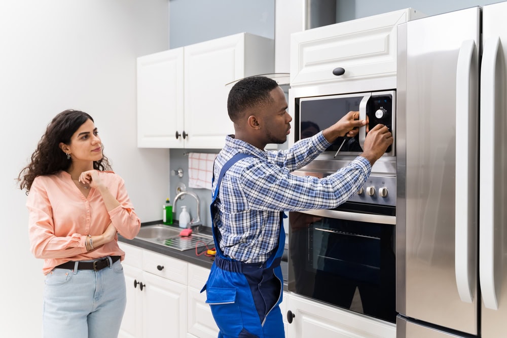 A repairman from MA appliance services in blue overalls fixes a built-in kitchen oven while a woman in a pink shirt watches thoughtfully beside him. The scene takes place in a modern white kitchen.