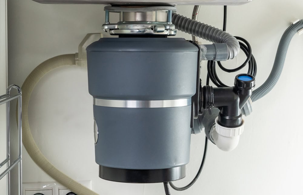A close-up view of a garbage disposal unit installed under a kitchen sink in Middlesex County, showing connected pipes, hoses, and wires against a white wall—ideal for those seeking same day appliance repair.
