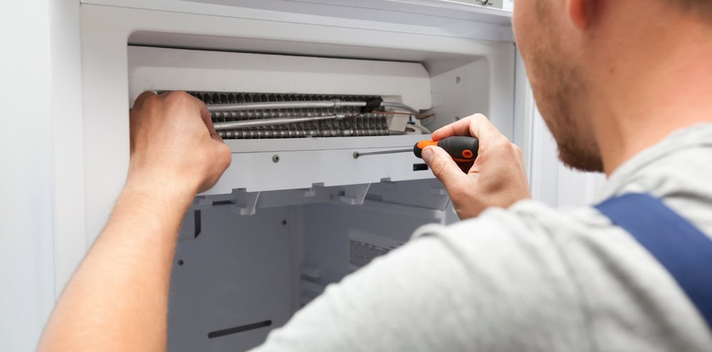 A person uses a screwdriver to repair or inspect the interior components of a refrigerator, focusing on the freezer section, demonstrating the quality of repairs for appliances offered by MA appliance services.