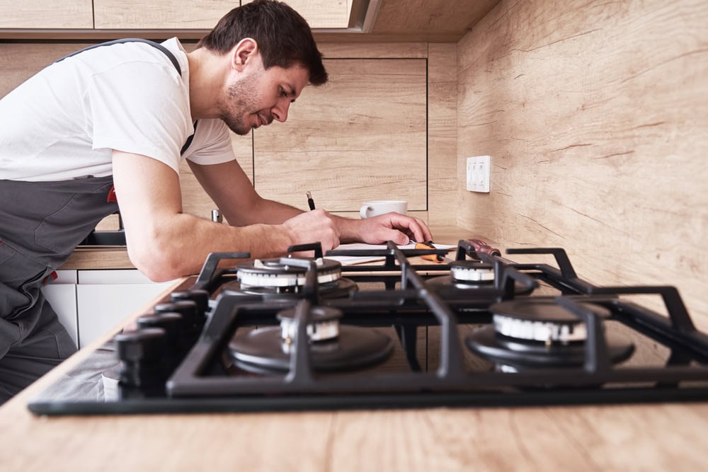 A man in work overalls, likely one of the skilled appliance technicians from MA Appliance Services, is writing on a clipboard in a kitchen with light wood cabinets and countertops, focusing on a modern gas stove.