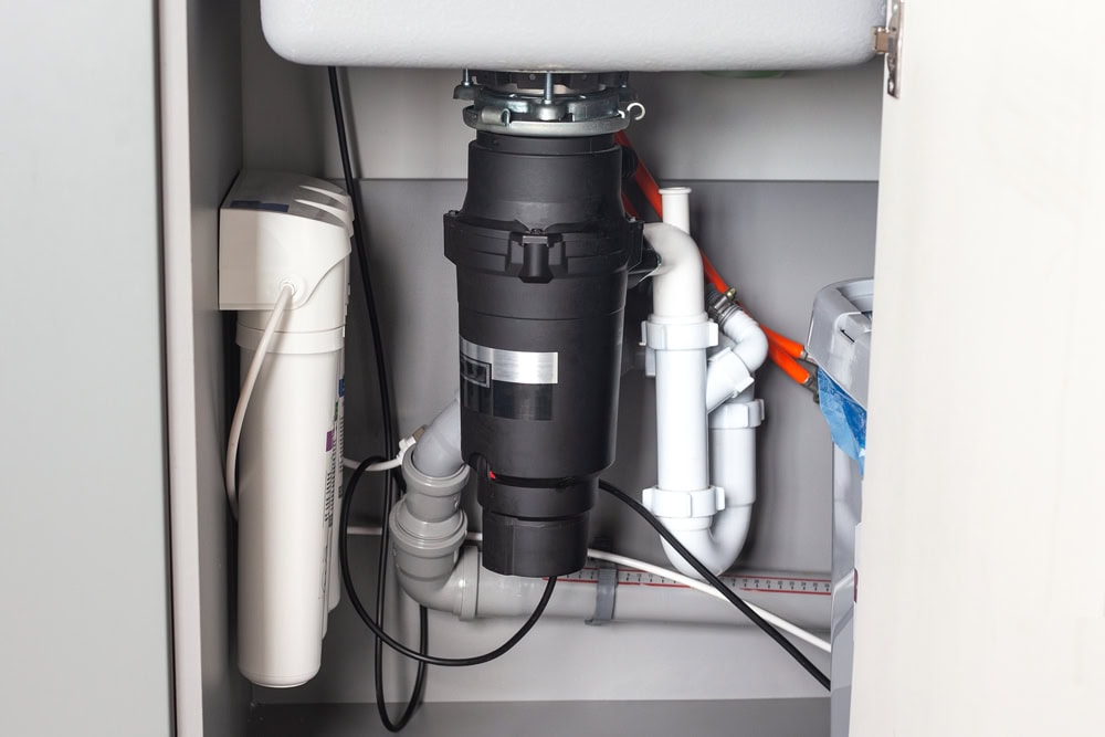 A close-up view of plumbing under a kitchen sink in Middlesex County, featuring a black garbage disposal unit, white pipes, water filter components, and various connections inside a cabinet.