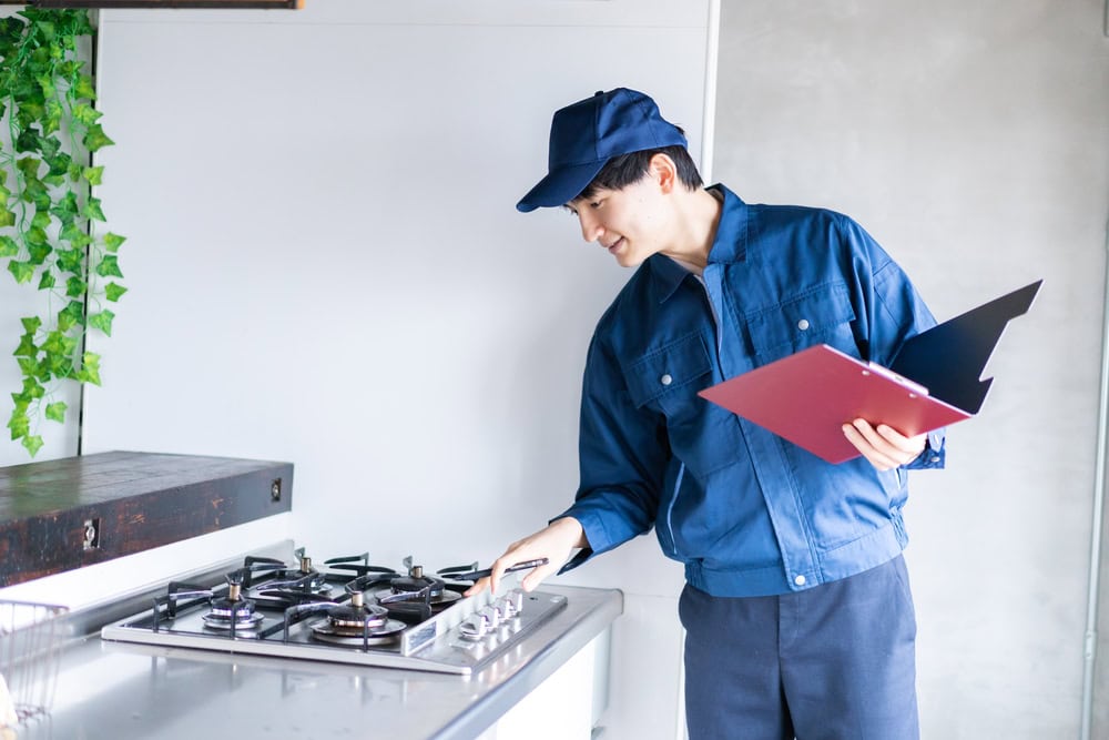 A technician in a blue uniform and cap inspects a kitchen gas stove while holding a clipboard, possibly checking for safety or functionality—typical of same day appliance repair by MA appliance services in Middlesex County.
