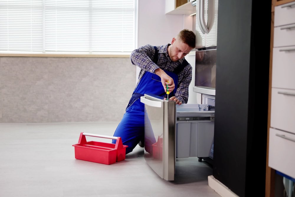 A repairman in blue overalls kneels on the floor, using a screwdriver to fix an open refrigerator, with a red toolbox beside him in a bright, modern kitchen—ideal for same day appliance repair in Middlesex County.