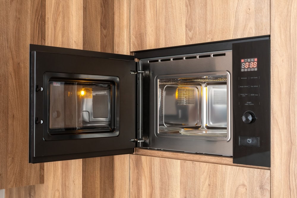A modern microwave oven with its door open, built into a wooden kitchen cabinet. The interior light is on, and the digital display shows numbers. Appliance technicians from Middlesex County, MA appliance services can keep it running smoothly.
