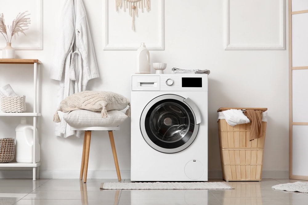 A modern laundry room with a white washing machine, a wooden stool stacked with folded towels and pillows, a wicker basket with clothes, and shelves with decor against a white wall—perfect for homes needing same day appliance repair.