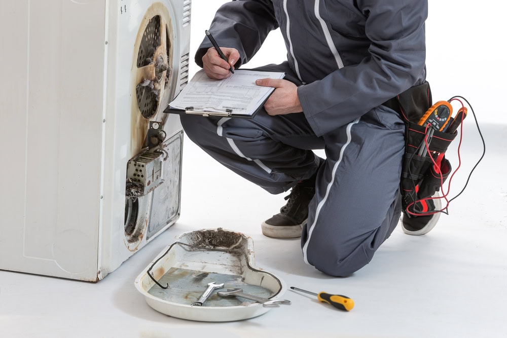 A repair technician in a gray jumpsuit is kneeling beside an open washing machine, taking notes on a clipboard. Tools and a removed machine panel are on the floor, showcasing expert MA appliance services for prompt repairs for appliances.