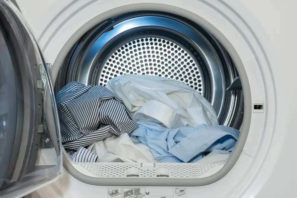 A front-loading washing machine with its door open, showing a pile of clean clothes inside, including white and blue shirts.