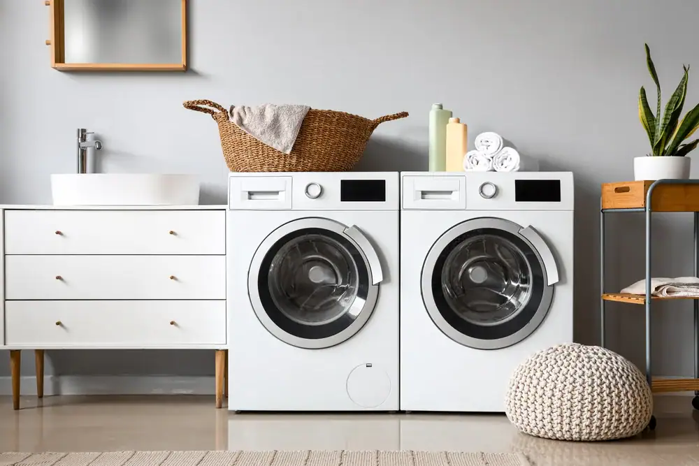 A modern laundry room with a front-loading washing machine and dryer side by side, a wicker basket on top, folded towels, and decorative items on nearby shelves, with neutral colored decor.