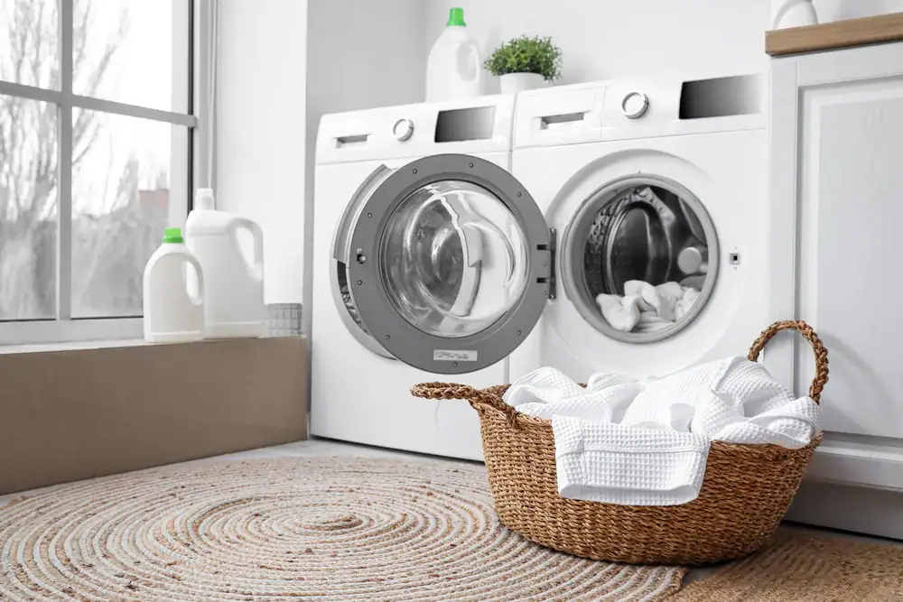 A laundry room with a washing machine and dryer, one door open, white towels in a wicker basket on a woven rug, and detergent bottles on the windowsill and countertop.