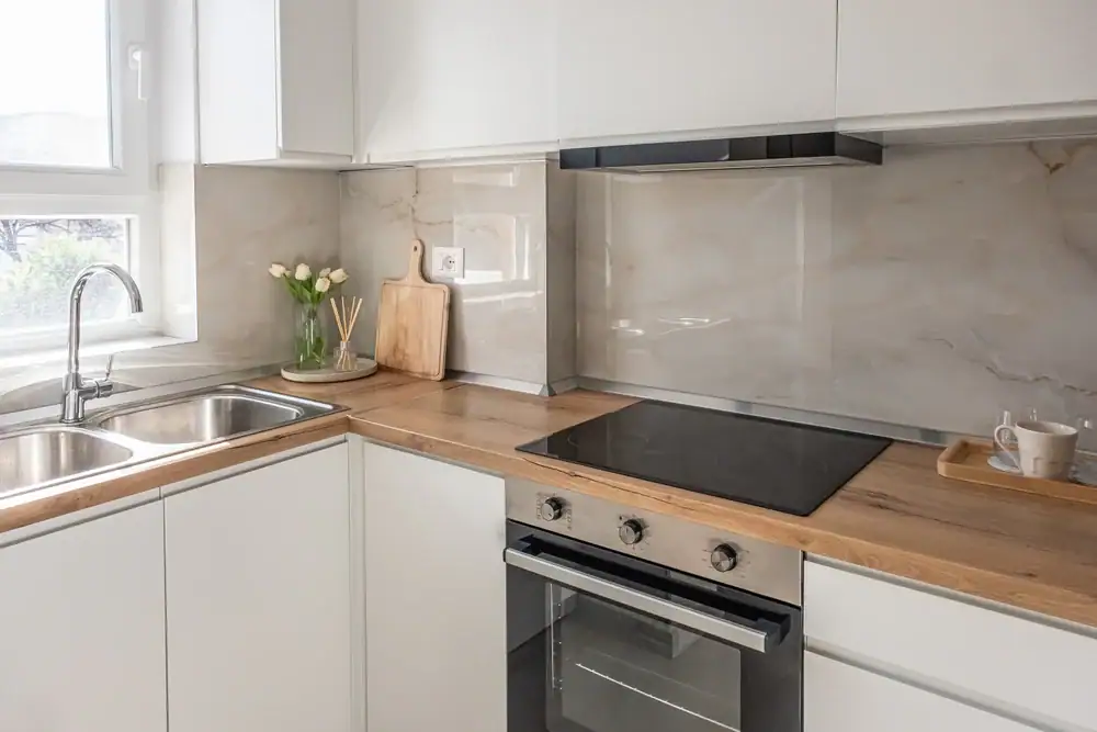 Modern kitchen with white cabinets, a wooden countertop, an electric stove, a sink by the window, and a marble backsplash. A cutting board, vase of flowers, and tray with teacups decorate the space.