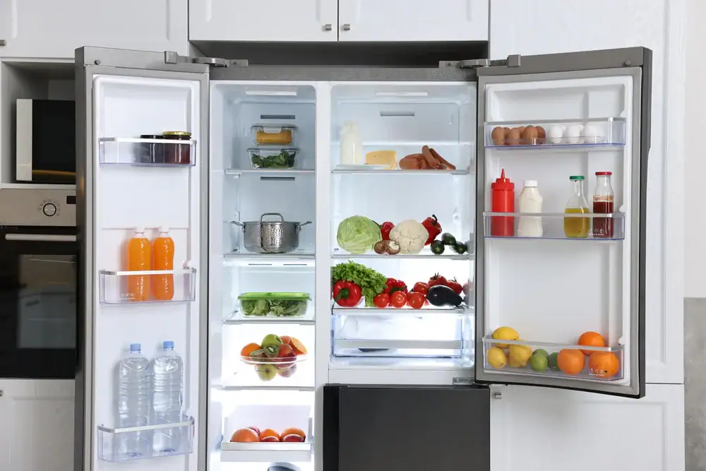 An open refrigerator filled with various foods, including vegetables, fruit, bottled drinks, eggs, sauces, and containers, neatly arranged on shelves and in the door compartments.