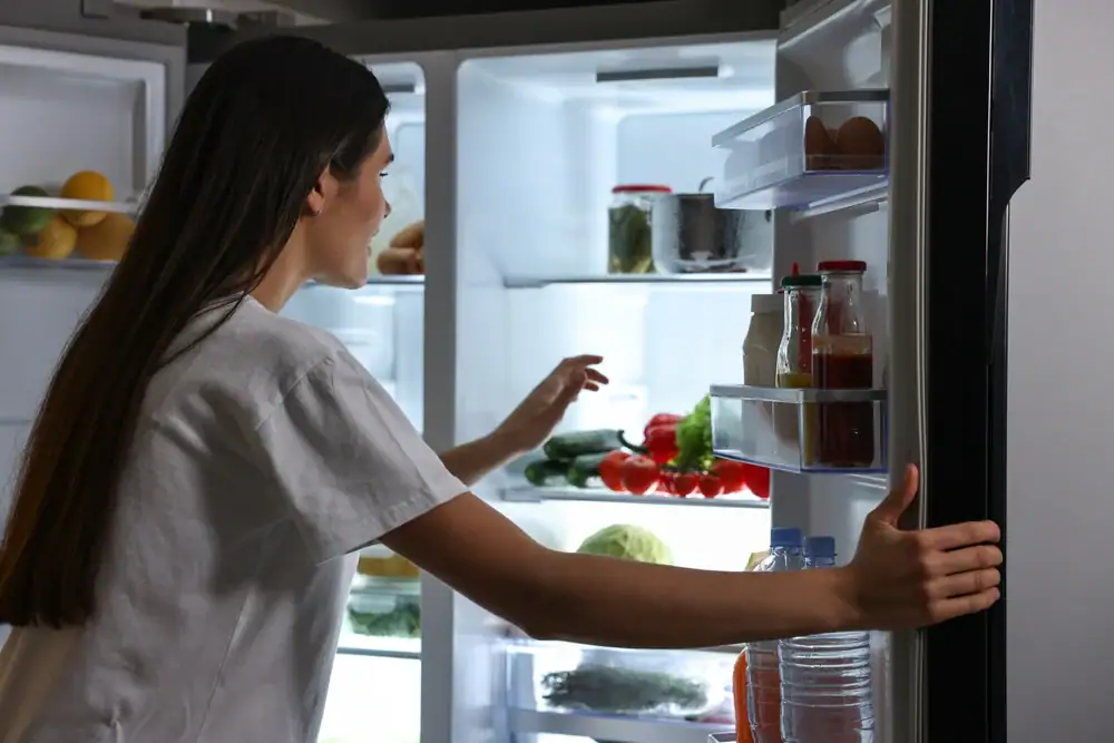 A woman with long dark hair wearing a white shirt opens a refrigerator filled with various food items, including vegetables, jars, bottles, and water bottles, while reaching inside.