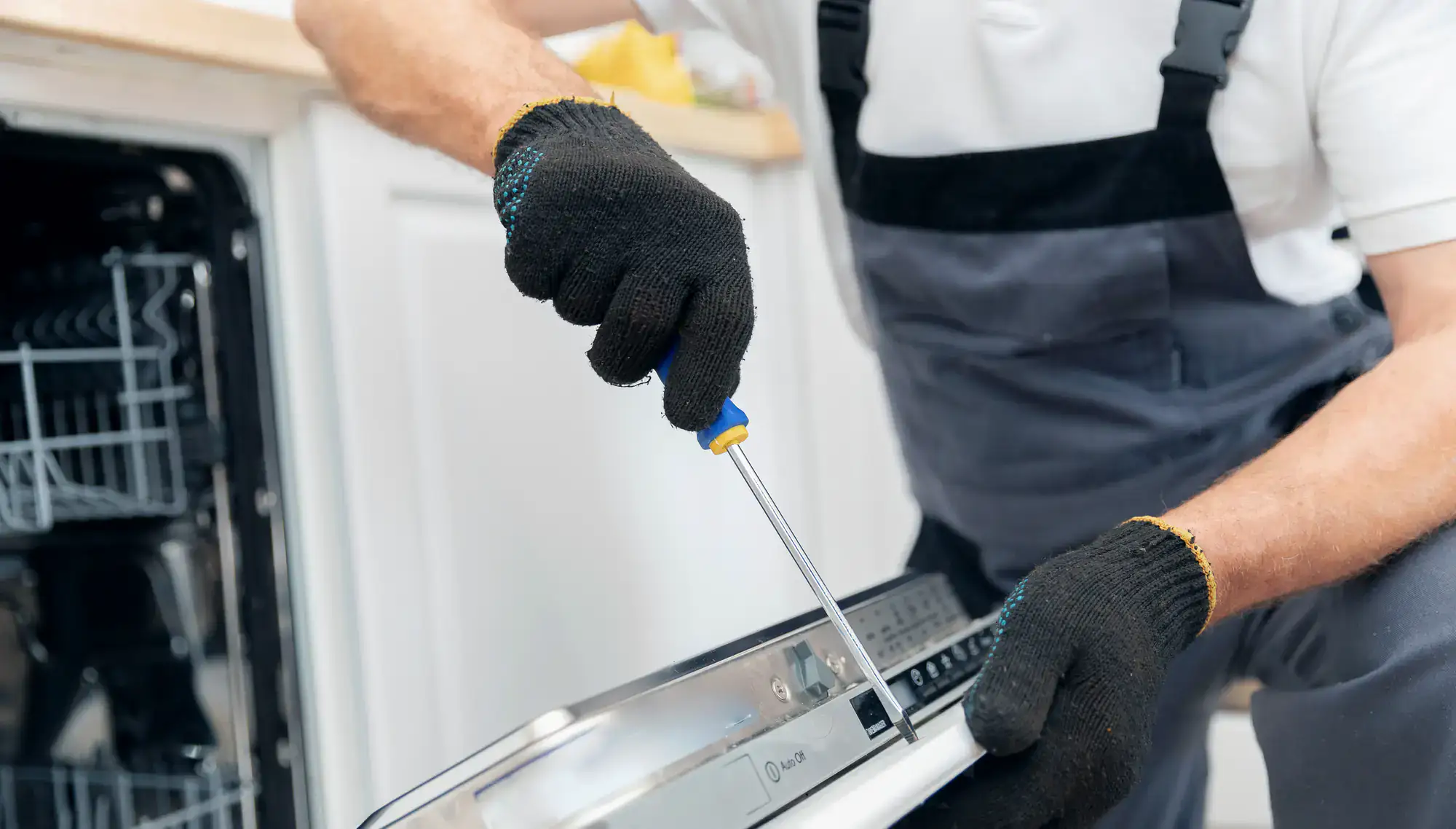 A person wearing black gloves uses a screwdriver to repair the control panel of a dishwasher, with the appliance door open and kitchen cabinets visible in the background.