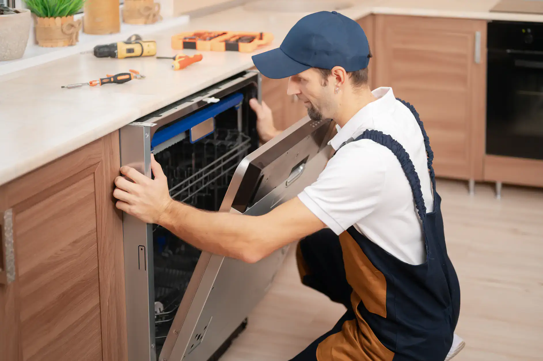 A repair technician in a uniform and cap kneels on the floor, inspecting the open door of a built-in dishwasher in a modern kitchen. Tools are placed on the countertop above.